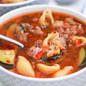 A white bowl filled with minestrone soup, featuring ground meat, shell pasta, zucchini slices, and chopped tomatoes in a broth. A spoon rests inside the bowl. A whole tomato and a striped cloth are visible in the background.