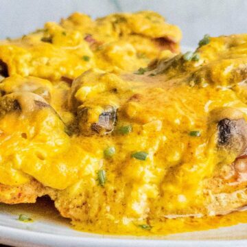 A close-up of a plate with chicken breasts smothered in a melted cheese and mushroom sauce. This low-carb dinner is garnished with chopped herbs and drizzled in a rich, yellow sauce, making it an enticing choice for those seeking delicious low-carb recipes.