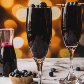 Two tall, elegant glasses filled with a dark beverage reminiscent of a classic kir royale sit on a marble surface. Blueberries garnish the rim of each glass. A small bowl of blueberries and a half-filled carafe are in the background, against a blurred, warm bokeh backdrop.