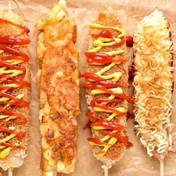Six skewered corn dogs lie on parchment paper. Each is covered in a variety of toppings such as sauces, cheese, and coatings like chopped potatoes and noodles. The sauces appear to be ketchup and mustard.