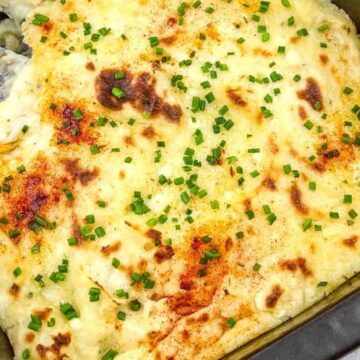 A baking dish filled with baked mashed potatoes topped with melted cheese, browned spots, and garnished with chopped chives. A serving spoon is placed in the dish, indicating a portion has been taken out.