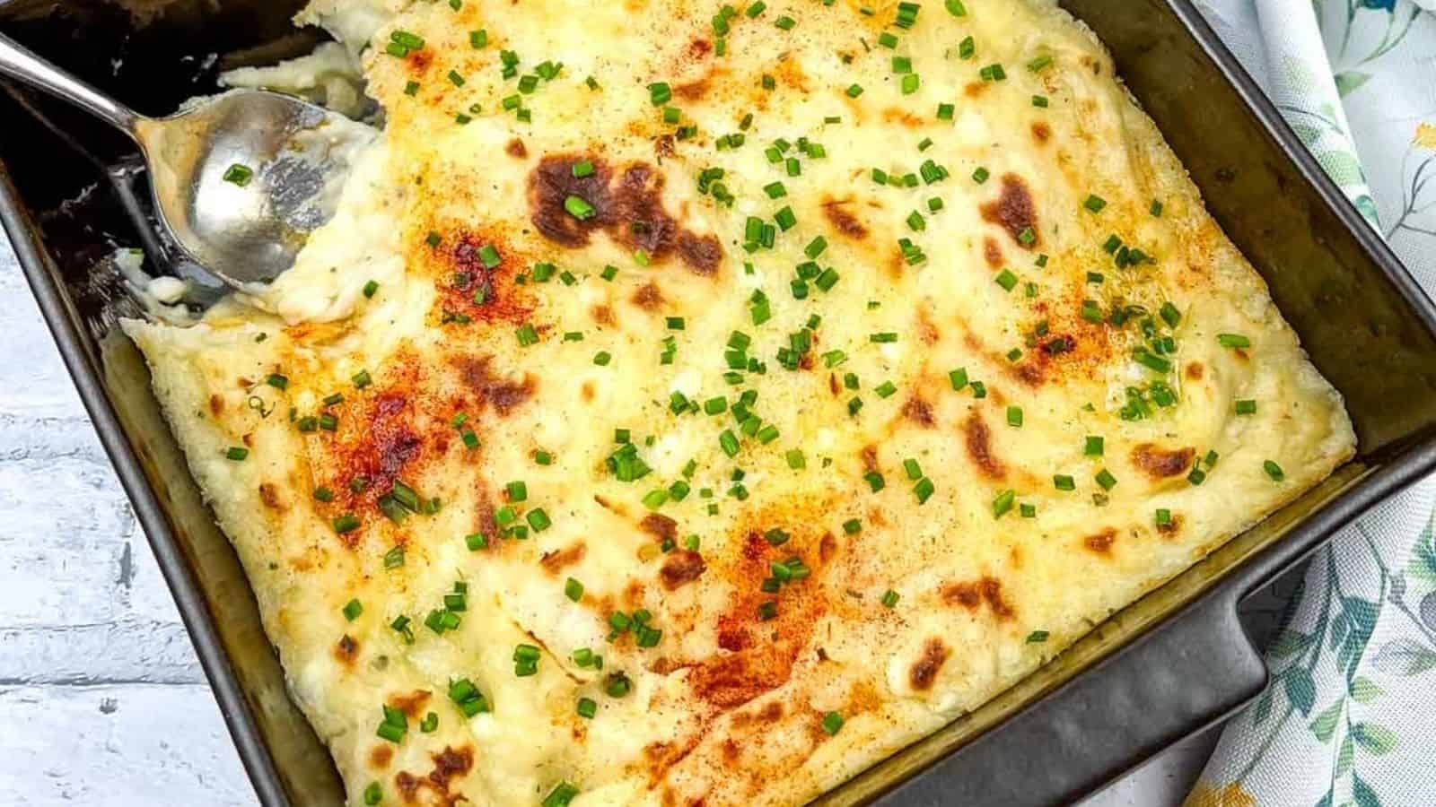 A baking dish filled with baked mashed potatoes topped with melted cheese, browned spots, and garnished with chopped chives. A serving spoon is placed in the dish, indicating a portion has been taken out.