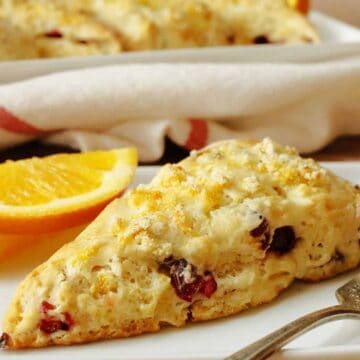 A triangular scone with visible chunks of cranberry on a white plate, accompanied by two slices of orange. A fork rests on the plate, and a striped cloth is in the background.