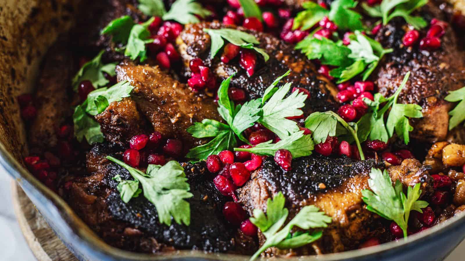 A close-up of a cooked dish featuring several pieces of meat topped with vibrant green parsley leaves and scattered pomegranate seeds. The dish is placed in a dark pot.