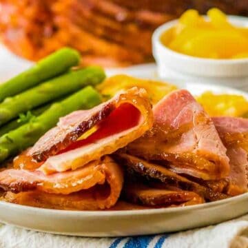 A plate of sliced ham served with cooked asparagus and a side of yellow fruit pieces, likely peaches, in a small bowl. The meal is set on a white towel with a blurred background of more food and a beverage.