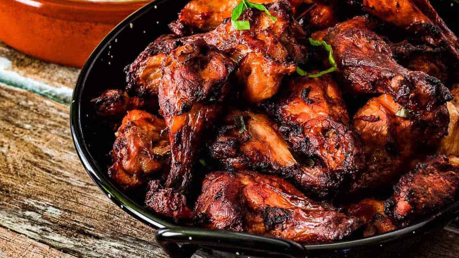 A black bowl filled with grilled chicken wings sits on a wooden surface, offering one of the world's most savory dishes. The wings are charred and garnished with a small sprig of fresh herbs, sparking culinary talk about flavor and presentation.