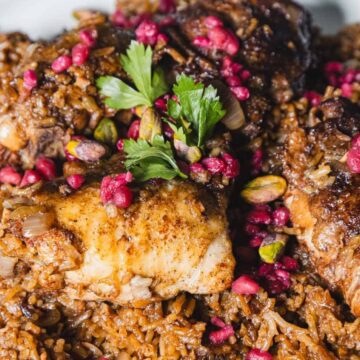 A close-up of a dish featuring seasoned rice topped with cooked chicken pieces. The meal is garnished with fresh parsley, pomegranate seeds, and pistachios, served on a white plate.