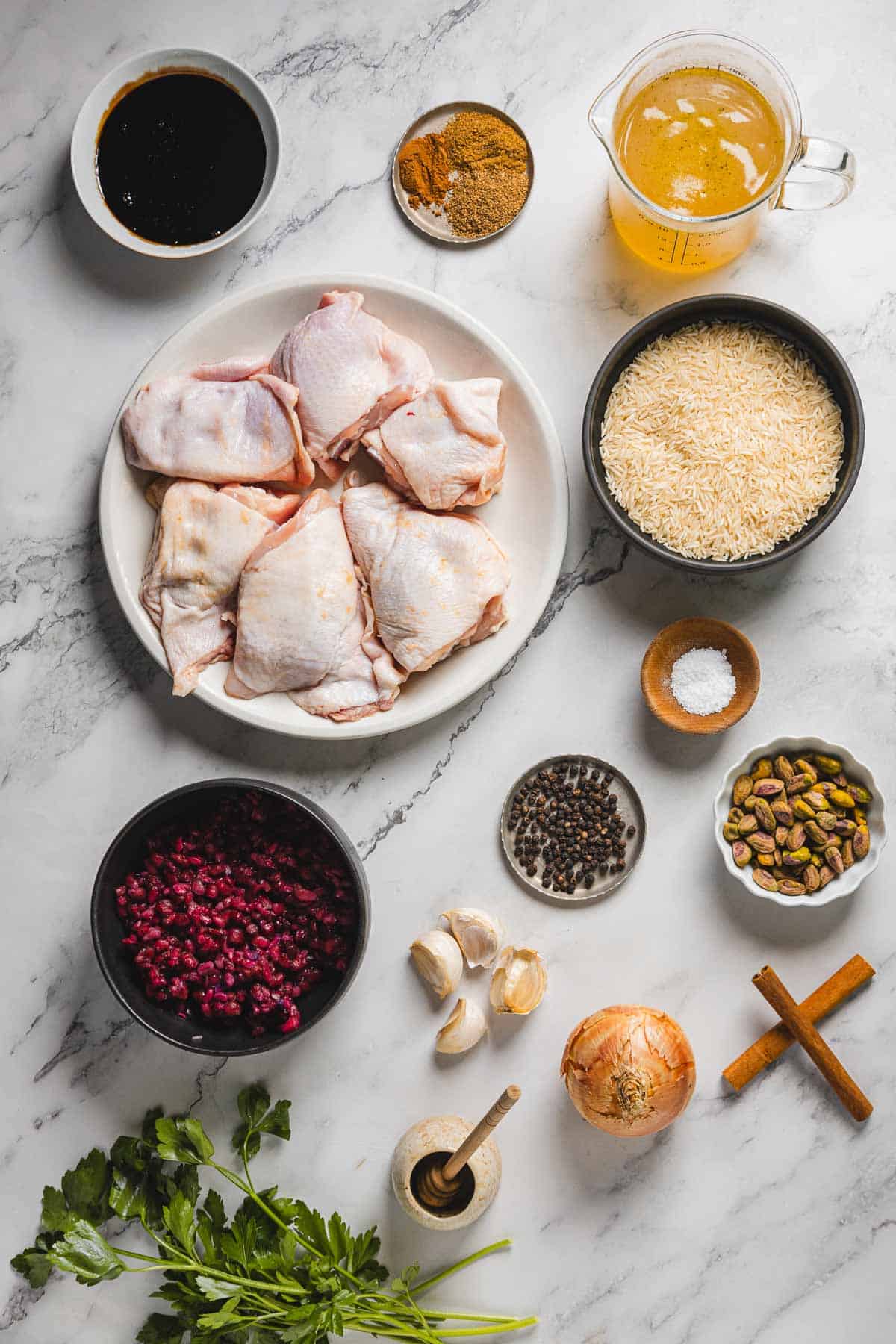 A selection of ingredients for pomegranate chicken and rice rests on a marble surface, showcasing raw chicken pieces, rice, vibrant pomegranate seeds, pistachios, garlic, an onion, fresh parsley, cinnamon sticks, peppercorns, salt, broth, soy sauce, and ground spices.