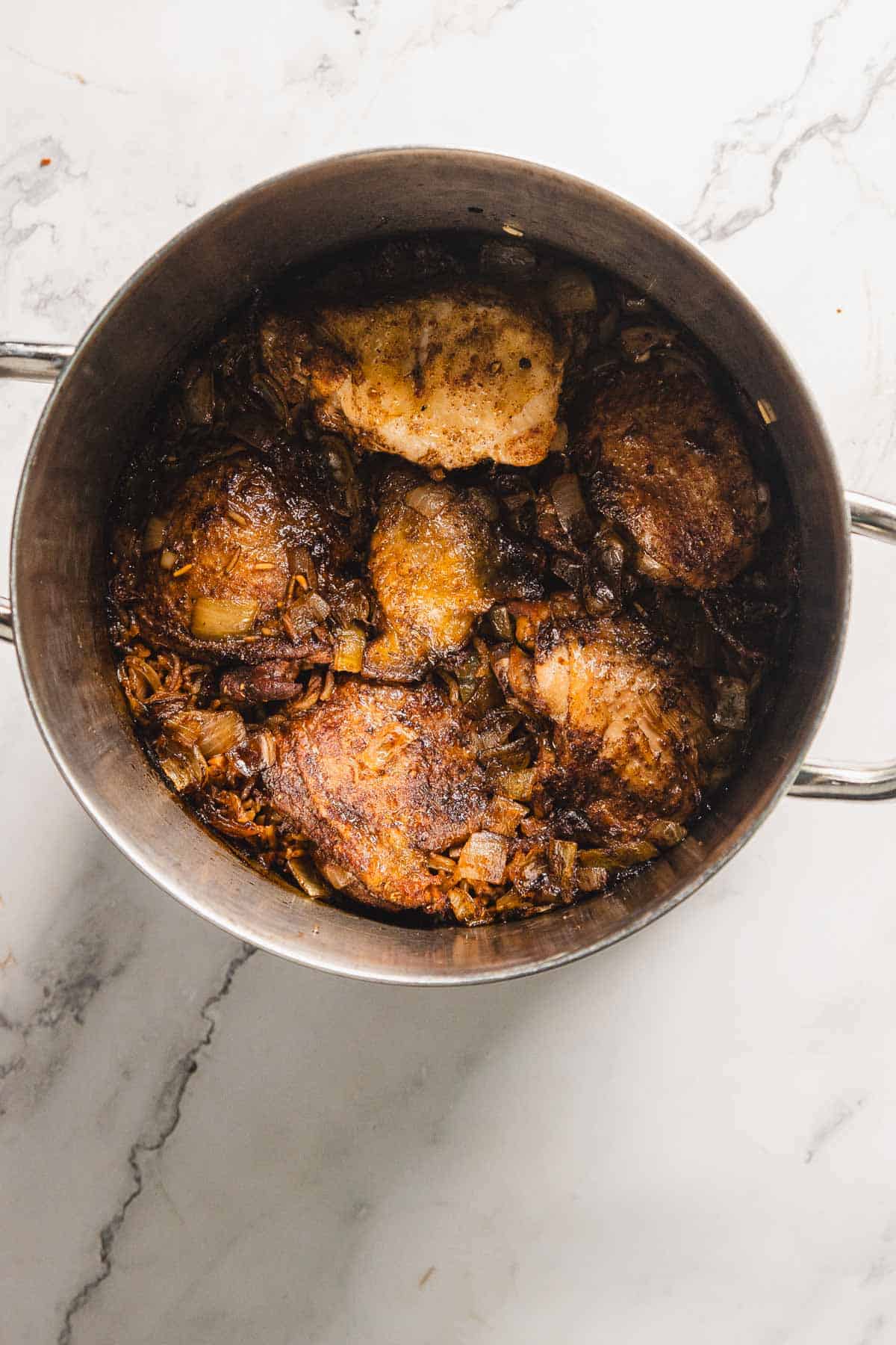 A pot containing pomegranate chicken pieces with browned onions on a white marble surface. The chicken appears seasoned and well-cooked, surrounded by caramelized onion pieces. The pot has two handles, hinting at the aromatic potential of rice to complete this savory dish.