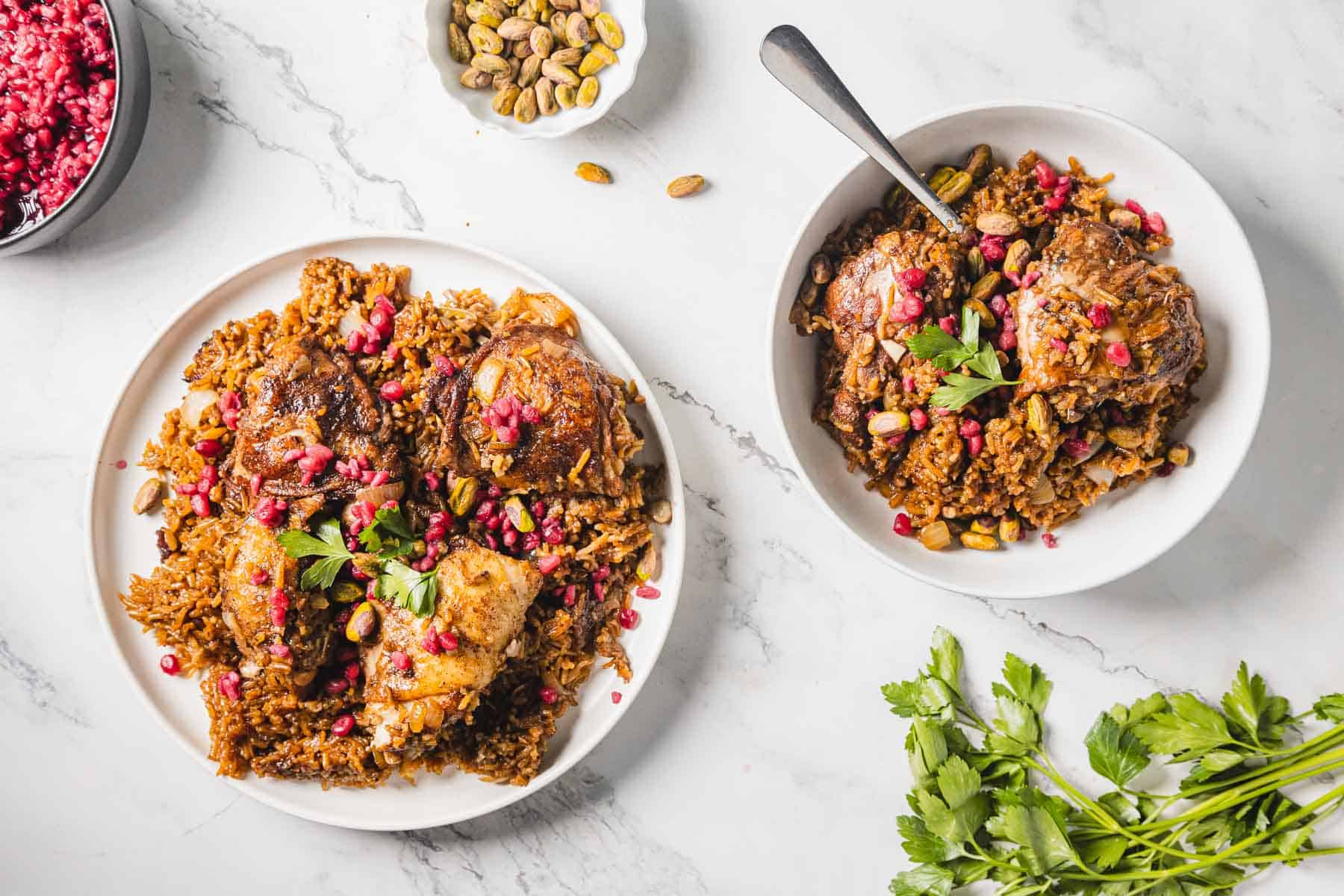 Two bowls of rice topped with succulent Pomegranate chicken, glistening pomegranate seeds, pistachios, and fresh parsley sit elegantly on a marble surface. Nearby, a small bowl of pistachios and a bunch of parsley add to this delightful 1-pot recipe's charm.