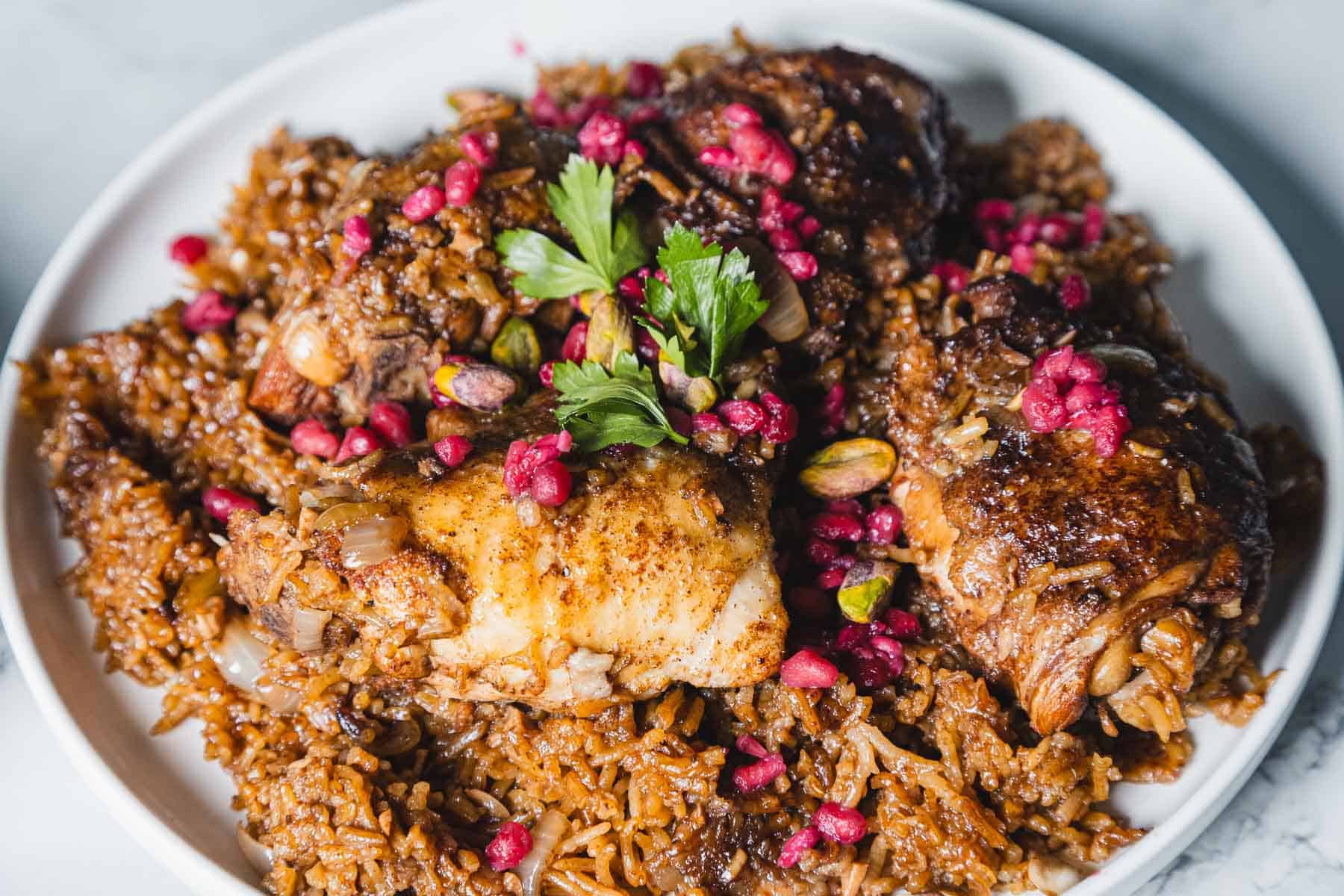 A delicious 1-pot dish of spiced rice mixed with tender pomegranate chicken, garnished with vibrant pomegranate seeds and a sprig of parsley. The meal is elegantly presented on a white plate, showcasing its rich brown hue from the spices.