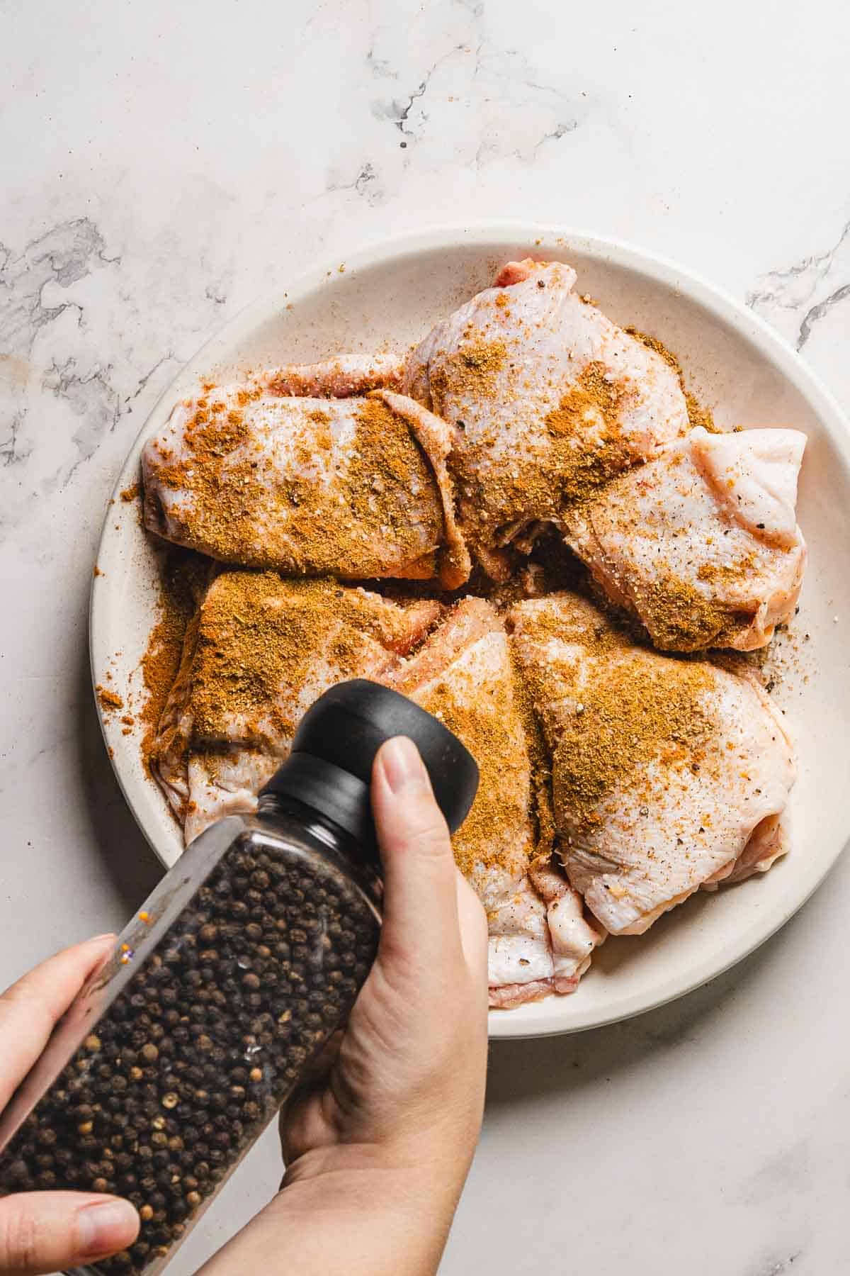 A person is holding a black pepper grinder over a white plate of raw, seasoned chicken thighs, ready to become a delightful pomegranate chicken and rice dish. The scene unfolds on a sleek marble countertop, with the hand poised in the bottom left corner of the image.