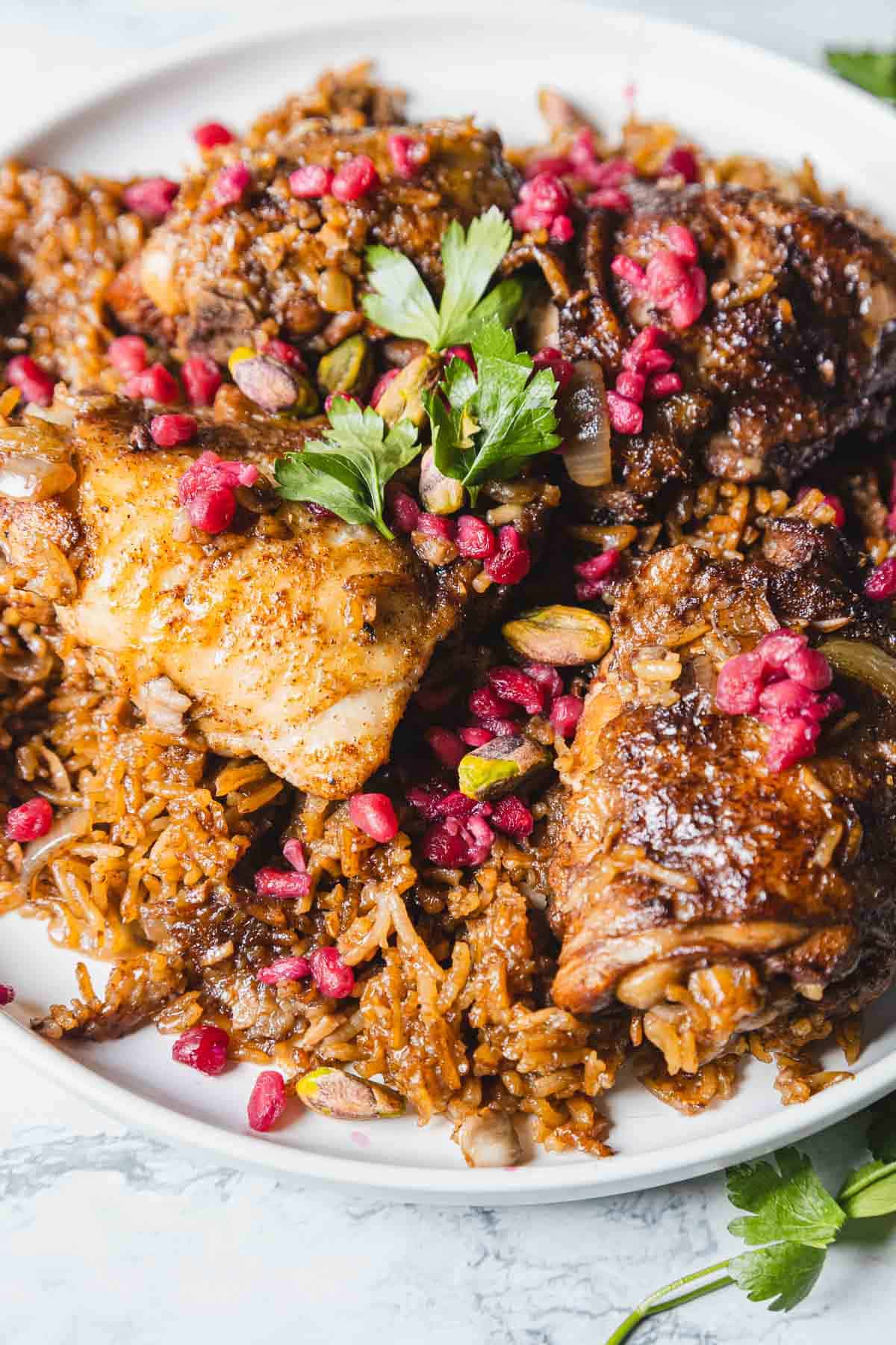 A delightful plate of 1-Pot Pomegranate Chicken features seasoned rice and three tender pieces of chicken adorned with pomegranate seeds. Garnished with parsley, the dish boasts visible nuts and herbs, elegantly presented on a round white plate.