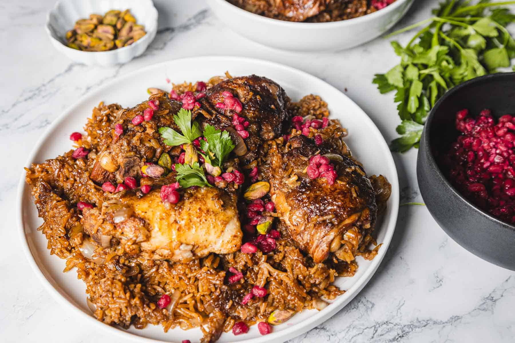 A dish of pomegranate chicken and rice is beautifully presented on a white plate, garnished with parsley and vibrant pomegranate seeds. Beside it, a small bowl of pistachios and a dark bowl with extra pomegranate seeds sit elegantly on the marble surface.