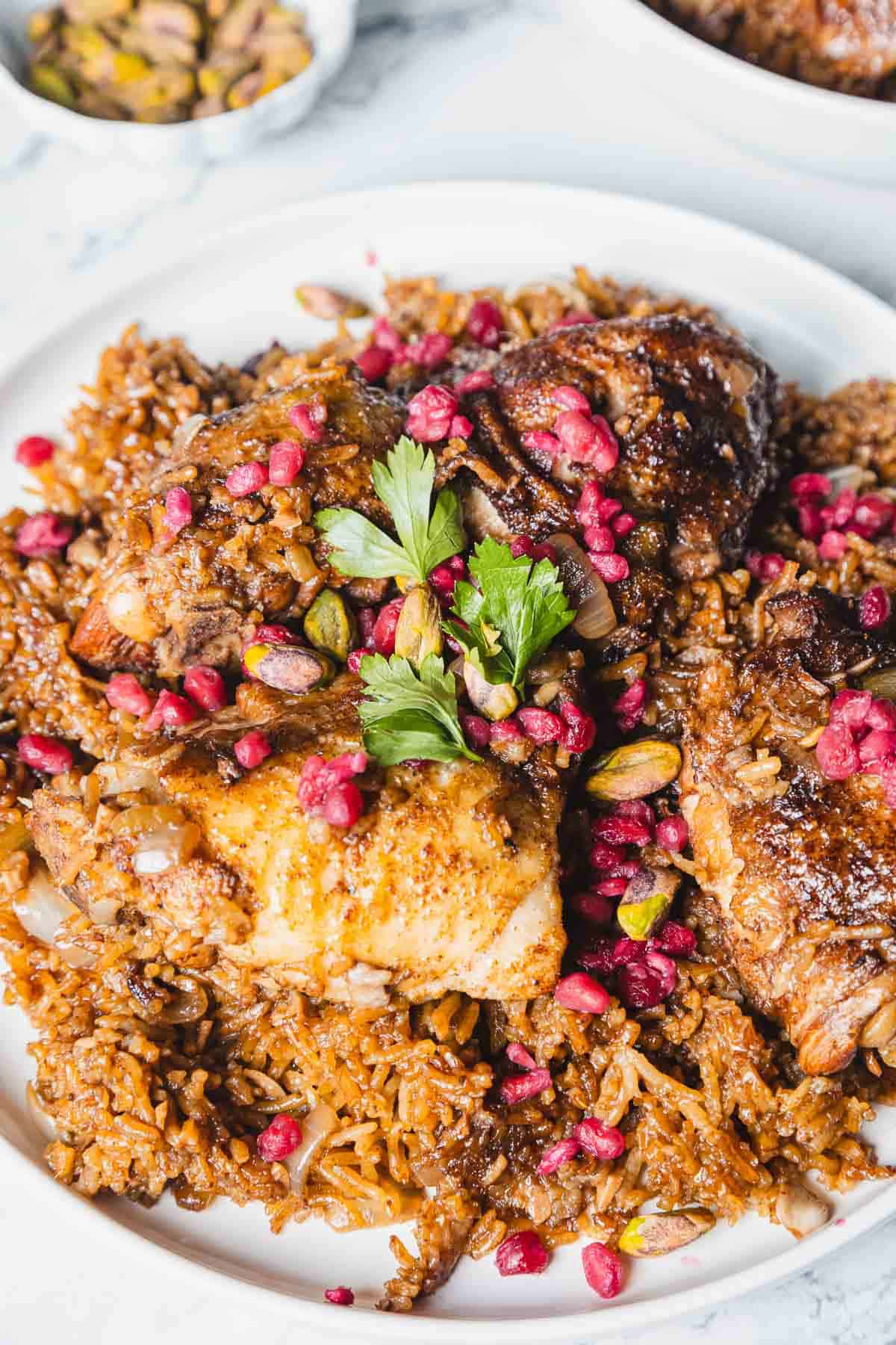 A white plate filled with seasoned pomegranate chicken and rice is topped with cooked chicken pieces. The dish is garnished with pomegranate seeds, nuts, and fresh parsley. In the background, a small bowl with more nuts is visible.