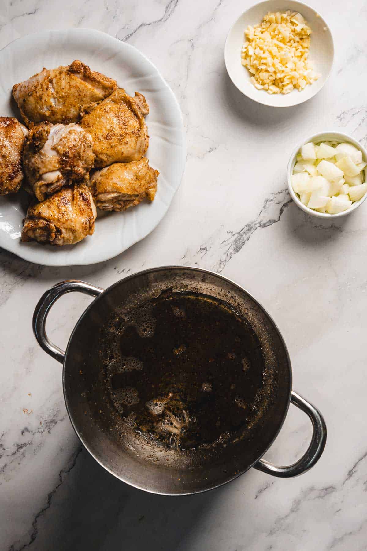 A frying pan with oil residue sits on a marble countertop. Nearby are a plate of juicy pomegranate chicken pieces, a small bowl of chopped onions, and another small bowl of minced garlic, ready to be combined with fluffy rice for a delightful meal.