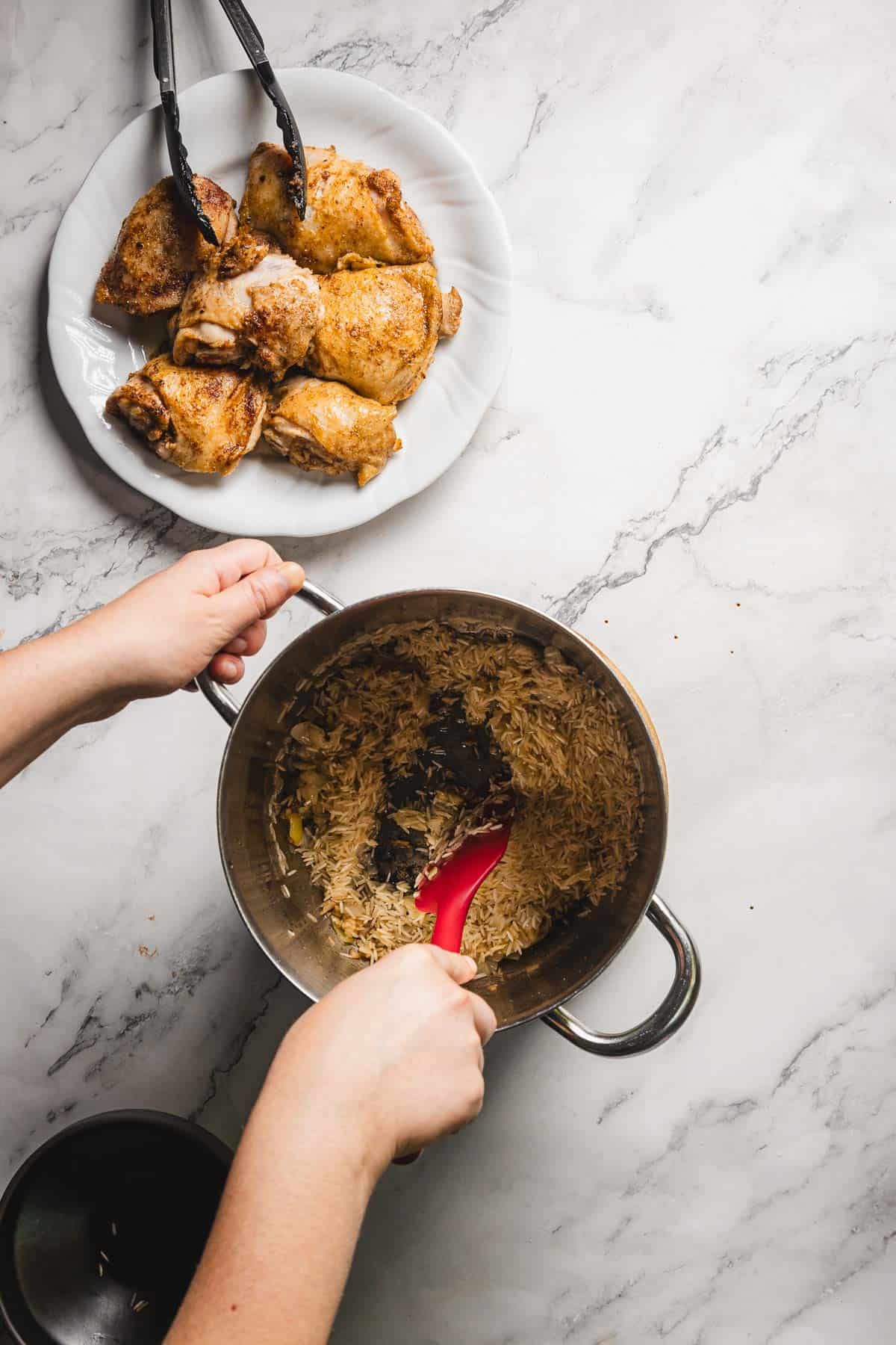 A person stirs browned rice in a saucepan with a red spatula, preparing the base for a flavorful pomegranate chicken and rice dish. Nearby on the marble countertop, a white plate holds cooked chicken pieces, while black tongs rest beside them, ready for use.