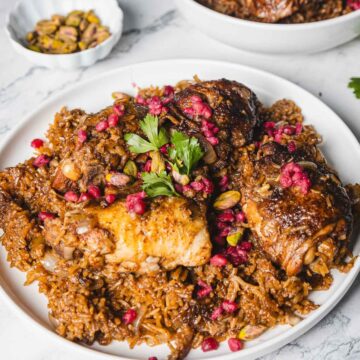 A plate of pomegranate chicken pilaf topped with vibrant seeds and garnished with parsley, this 1-pot recipe includes flavorful rice and pistachios. In the background, a small bowl brims with extra pistachios, complementing the dish.