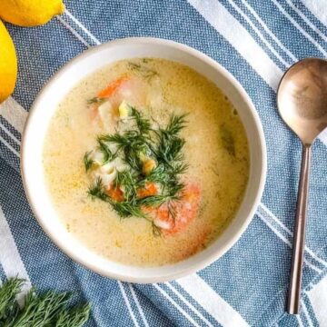 A bowl of creamy soup with herbs, carrot slices, and visible chunks of other vegetables sits on a blue and white striped cloth. A spoon and two whole lemons nestle beside the bowl. Perfect to pair with a blanket for cozy evenings, this dish warms both stomach and soul.