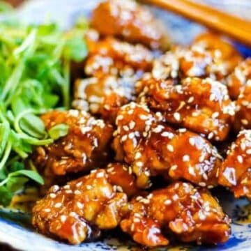 A plate of sticky honey garlic chicken wings topped with sesame seeds. Fresh greens are placed on the side. The dish is presented on a blue and white patterned ceramic plate. Chopsticks rest on the edge.
