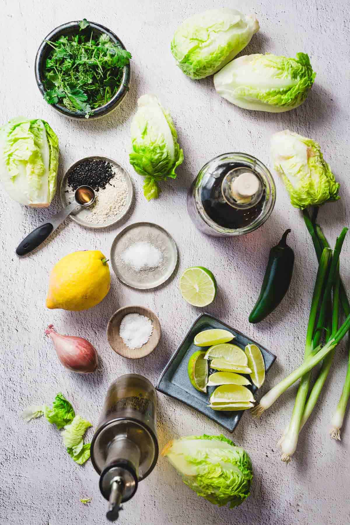 Top-down view of assorted ingredients on a light surface, including romaine lettuce heads, a lemon, lime wedges, a jalapeño, green onions, parsley, shallot, and various bowls containing black sesame seeds, seasonings, and oils.