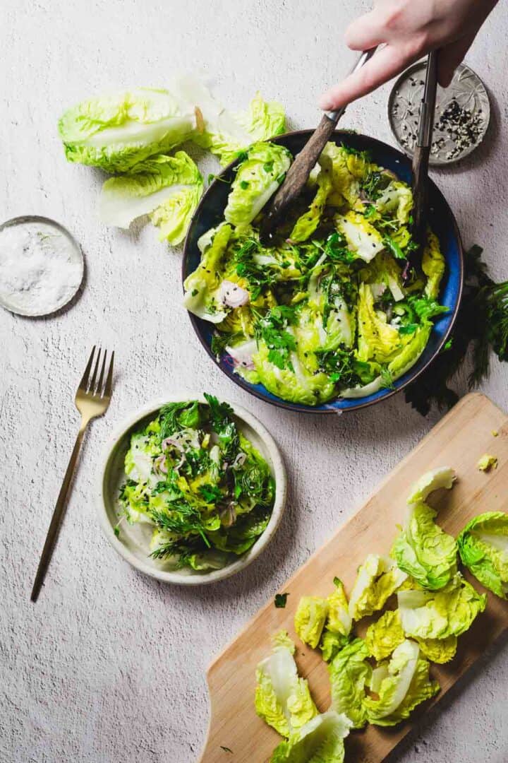 A person tosses a green salad with tongs in a large bowl on a white surface. A smaller bowl of salad with a fork is nearby. Lettuce leaves and herbs are on a cutting board, and a small dish of salt is visible.