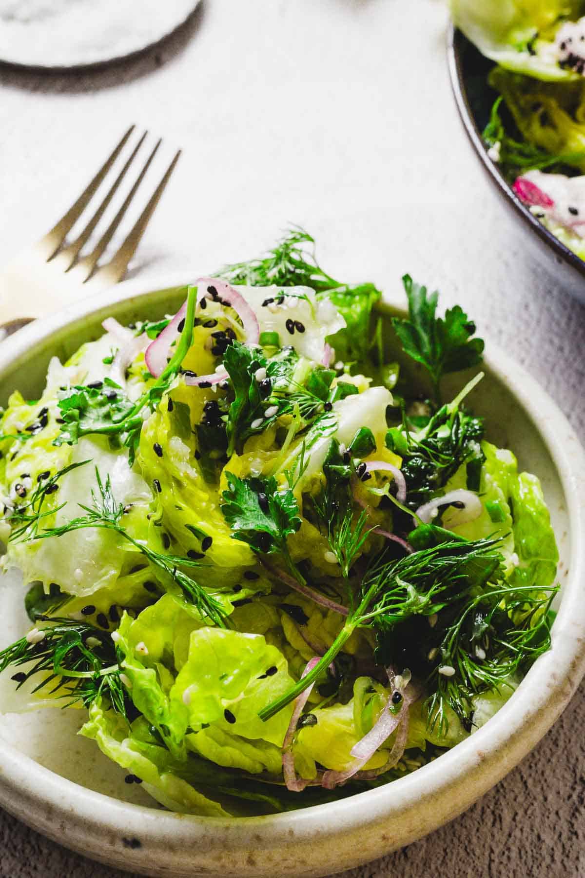 A bowl of fresh green salad featuring lettuce, dill, parsley, and sliced red onions, garnished with black sesame seeds. A fork rests on the table beside the bowl.