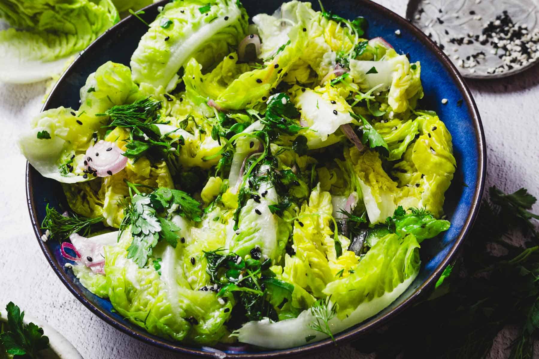 A close-up of a salad in a blue bowl. The salad consists of fresh lettuce leaves, garnished with herbs like parsley and dill, thinly sliced onions, and sprinkled with sesame seeds. The background shows additional salad ingredients.