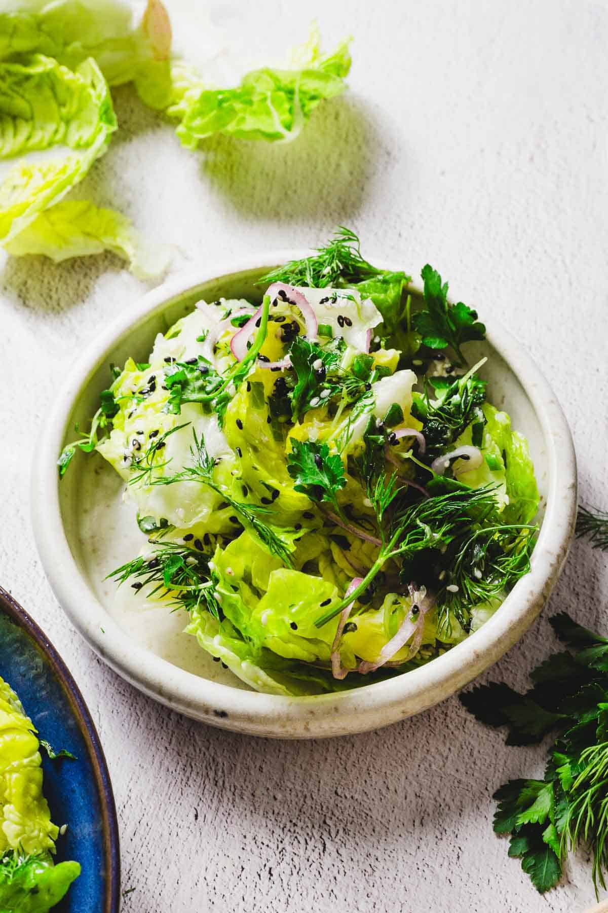 A bowl of salad with leafy greens, sliced onions, and fresh herbs, sprinkled with black sesame seeds. The background features additional lettuce leaves and herbs on a light-textured surface.