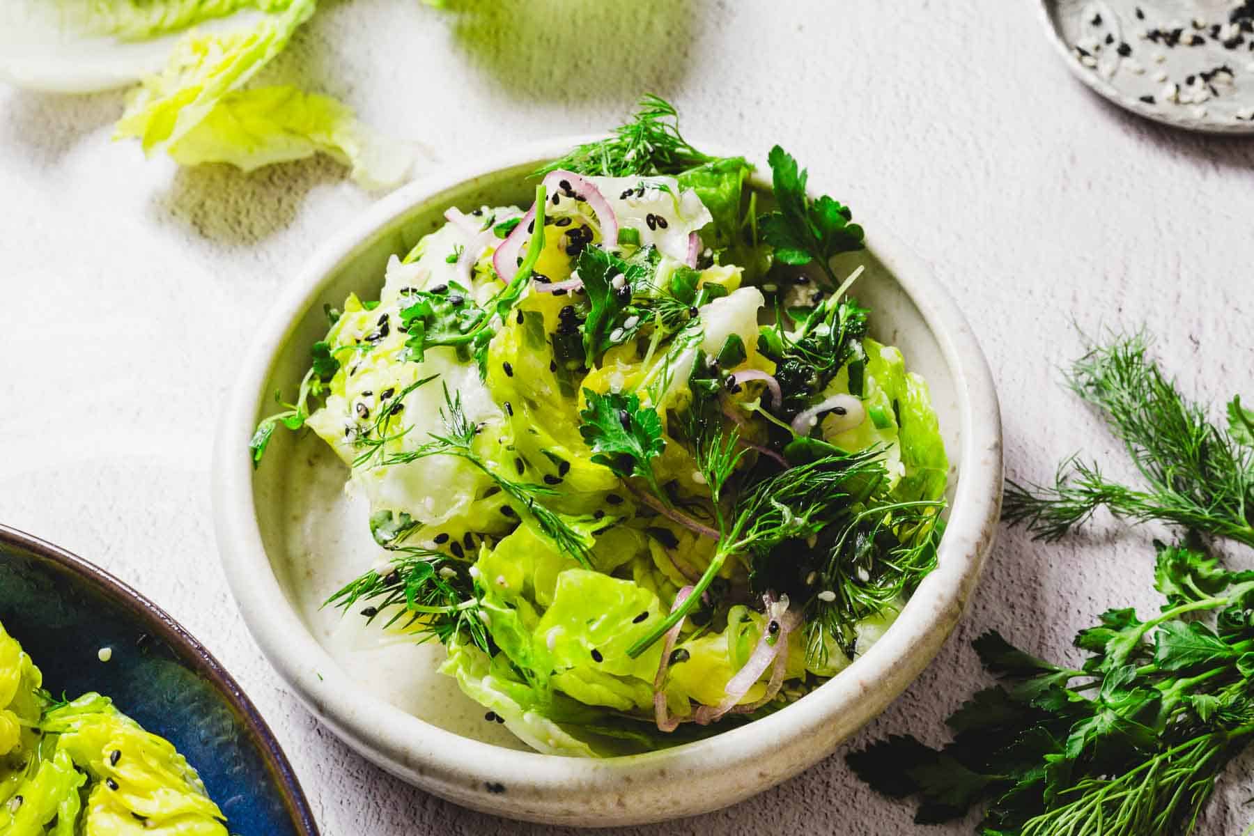 A bowl of fresh green salad with lettuce, dill, parsley, and sliced onions, garnished with black sesame seeds. Set against a light-colored textured surface with scattered herbs around.