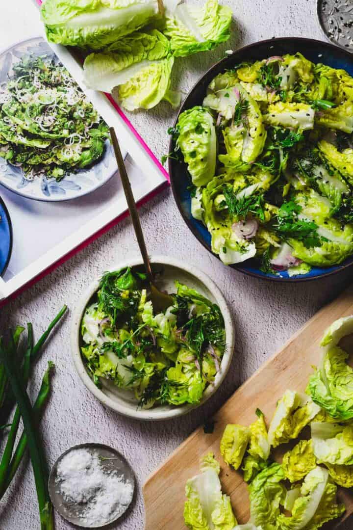 A table displays a fresh little gem salad with chopped lettuce, herbs, and sliced onions in multiple bowls. A cookbook page showing a similar salad is open nearby. Green onions and a small dish of salt are also visible on the table.