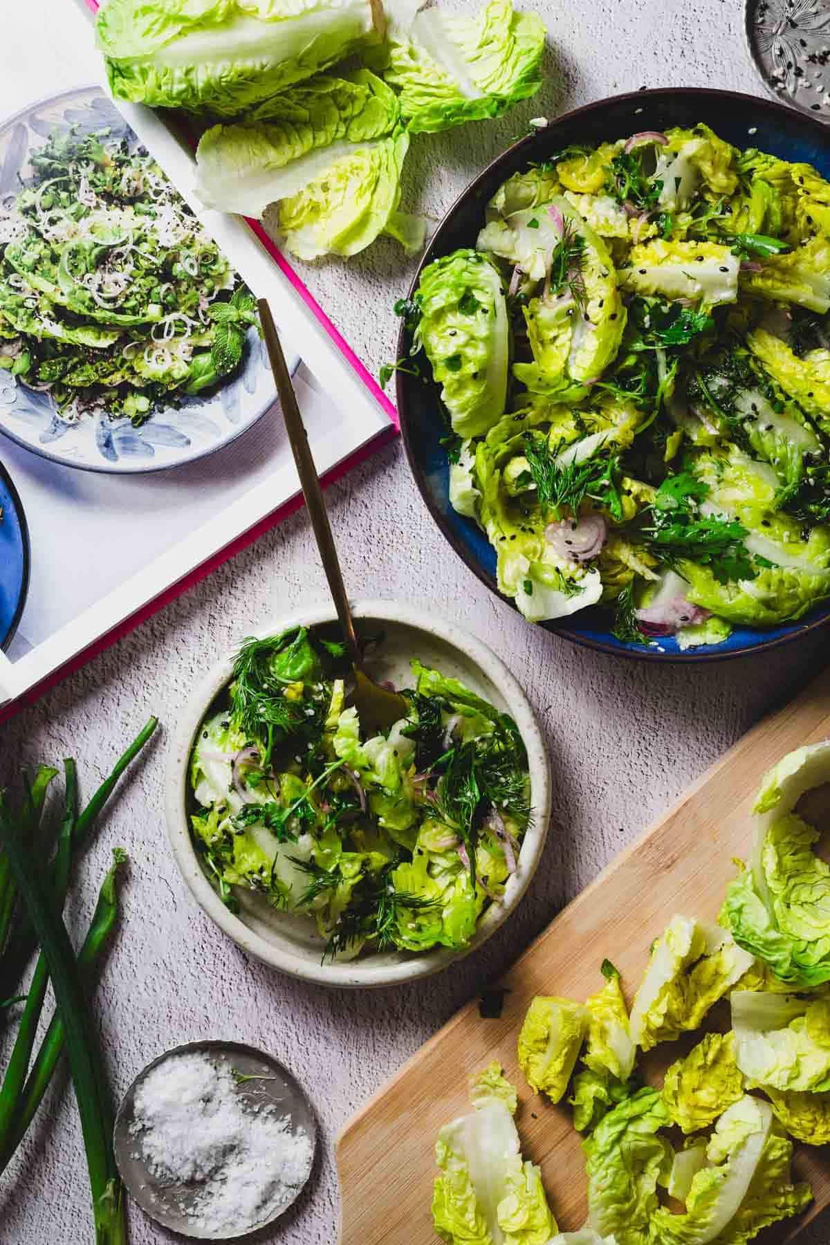 A variety of green salads featuring lettuce, fresh herbs, and sliced onions are displayed. A cookbook with a similar salad recipe is open in the background. Ingredients like salt and green onions are placed around the dishes on a light surface.