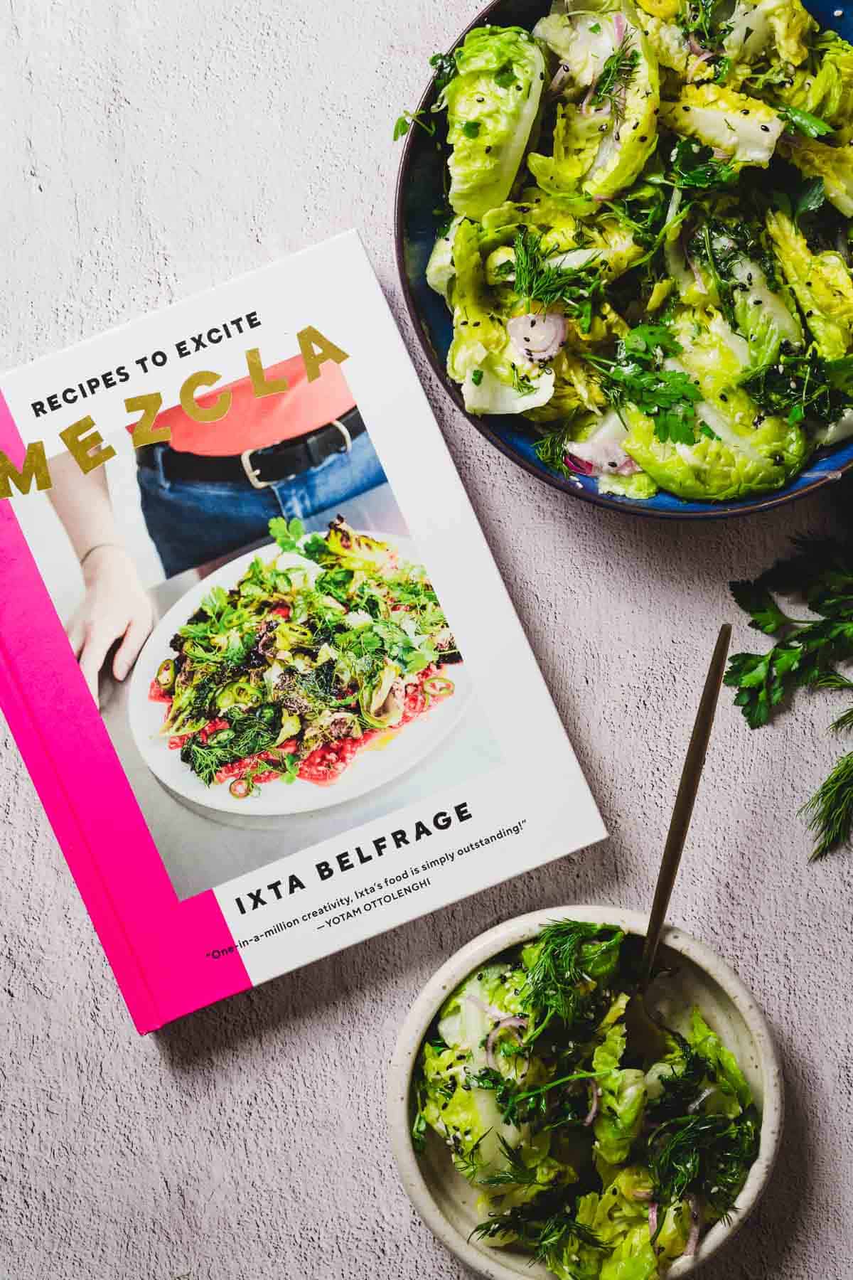 A cookbook titled "Mezcla: Recipes to Excite" by Ixta Belfrage is open on a table, next to a plate and a small bowl both filled with a salad featuring green leaves and herbs.