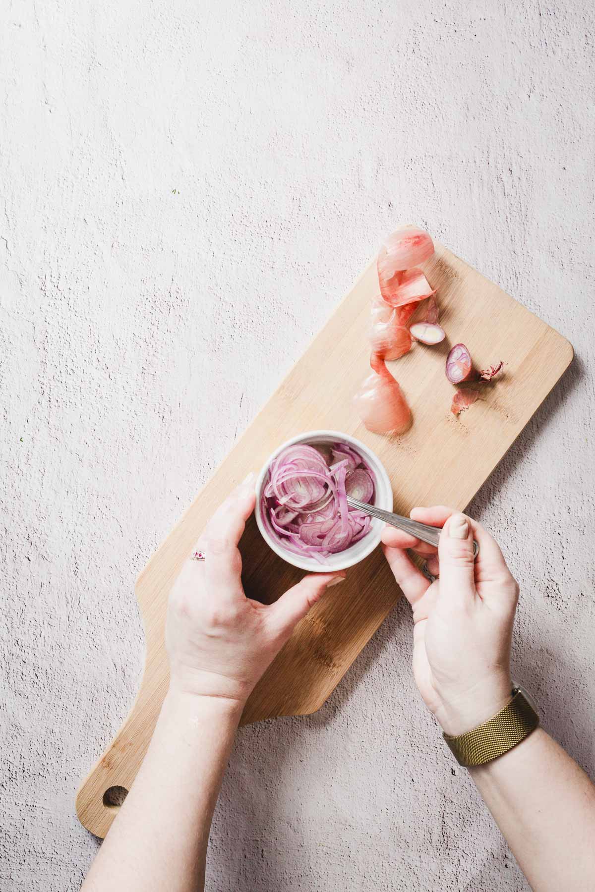 Hands are seen mixing sliced red onions in a small bowl with a spoon on a wooden cutting board. Additional onion slices and peels are scattered on the board. The background is a textured white surface.