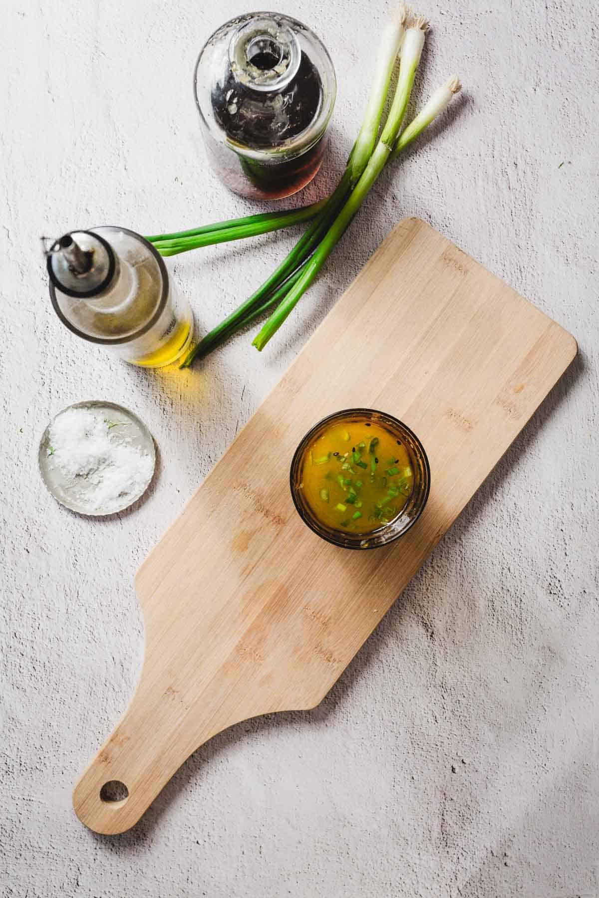 A wooden cutting board with a small bowl of yellow dressing garnished with chopped herbs. Nearby are two bottles, one with a dark liquid and the other with oil, a small dish of salt, and a few green onions on a textured surface.