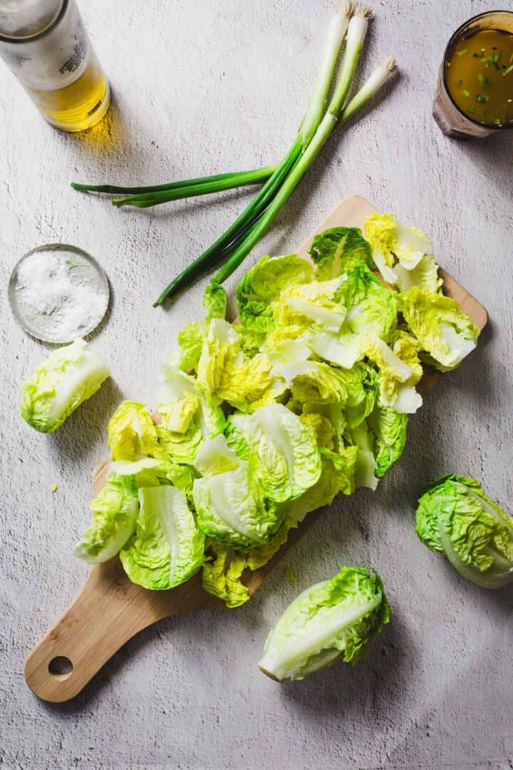 Chopped romaine lettuce on a wooden cutting board. Green onions are placed on the board. Nearby are a glass of beer, a small bowl of salt, and a glass with a light-colored drink garnished with herbs.