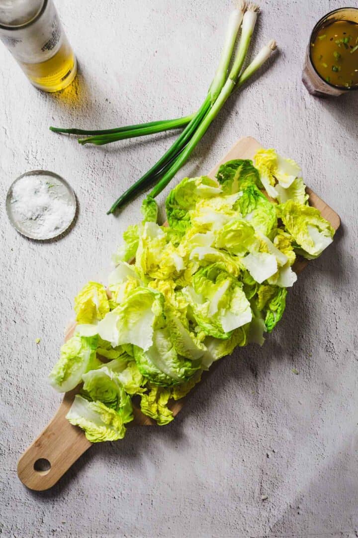 A wooden cutting board with chopped lettuce leaves, alongside green onions, a bowl of salt, a glass of amber liquid, and a jar of dressing on a light gray surface.