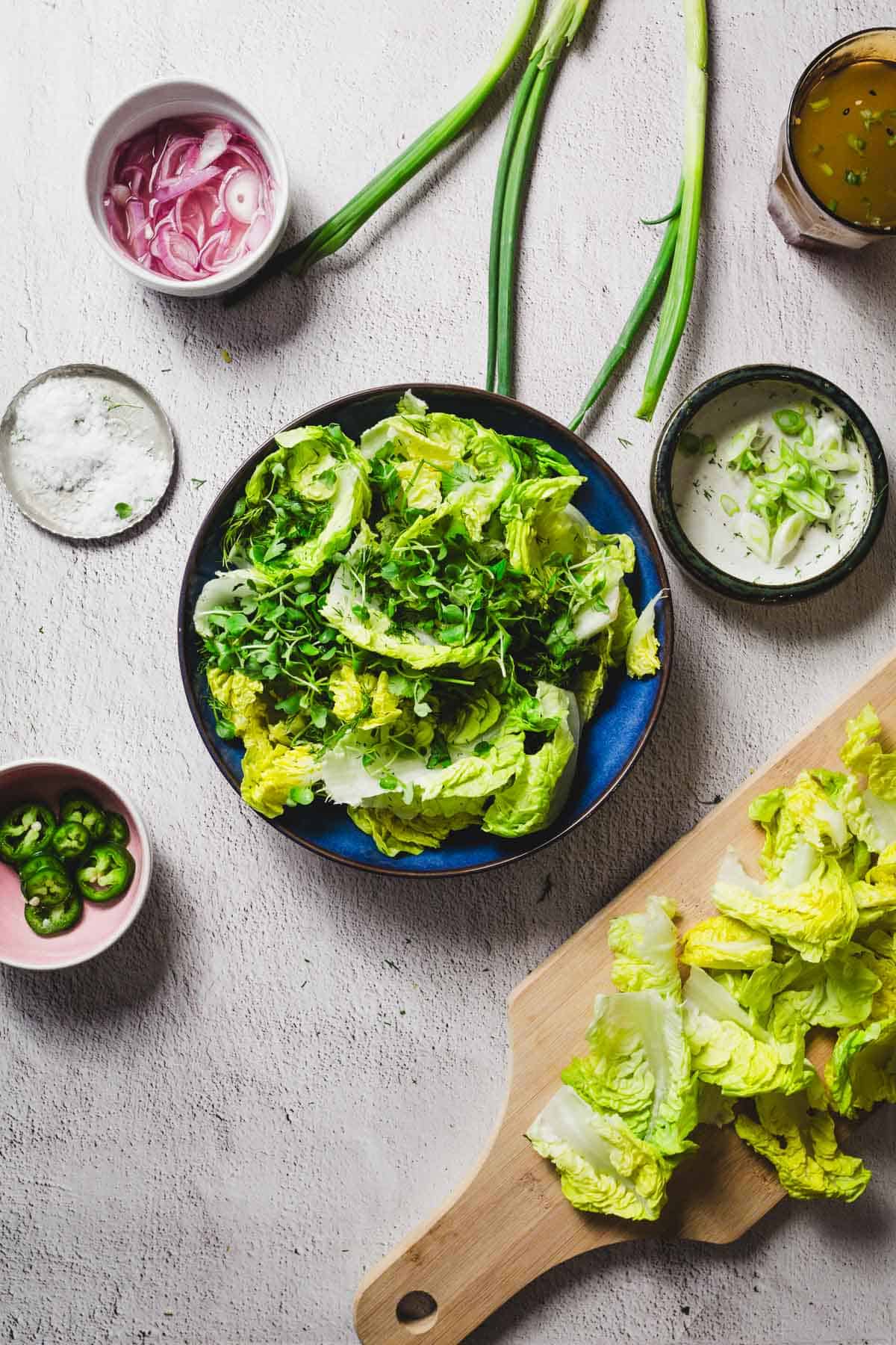 A top-down view of a table showcases a little gem salad masterpiece with a bowl of fresh lettuce and herbs. A cutting board holds crisp leaves, while small bowls brim with sliced onions, jalapeños, creamy dressing, and salt. Nearby, green onions stand ready for their garnish role.