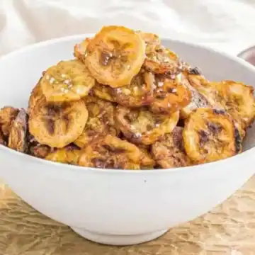 A white bowl filled with fried plantain slices is placed on a piece of parchment paper, perfect for whipping up anytime you crave low-cost snacks. There's a bottle of oil and a wooden bowl with salt in the background, set atop a rustic wooden surface.