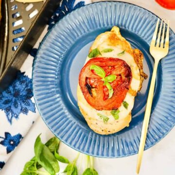A grilled chicken breast topped with sliced tomatoes and chopped basil on a blue plate with a gold fork. A decorative napkin with a floral design and a sprig of basil are nearby, with a glimpse of more chicken in the background.
