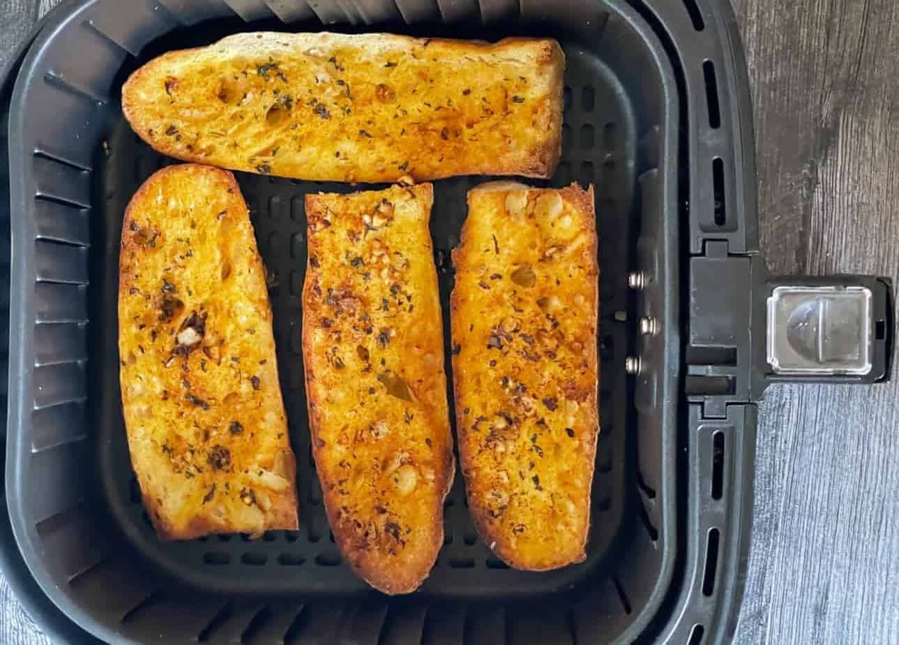 Four pieces of delicious toasted garlic bread are arranged inside an air fryer basket. Each slice is golden brown, with visible herbs and garlic on top, set against a dark, textured surface—perfect for exploring new bread recipes.