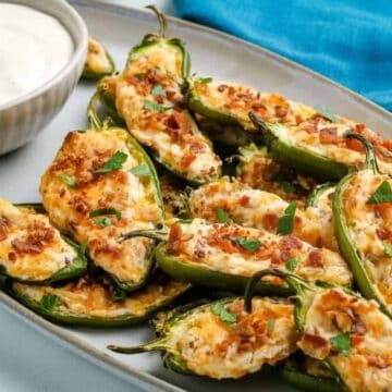 A plate of stuffed jalapeños filled with cheese and garnished with bacon bits and herbs. A small bowl of white dipping sauce sits beside them on a gray oval platter. A blue cloth is in the background.