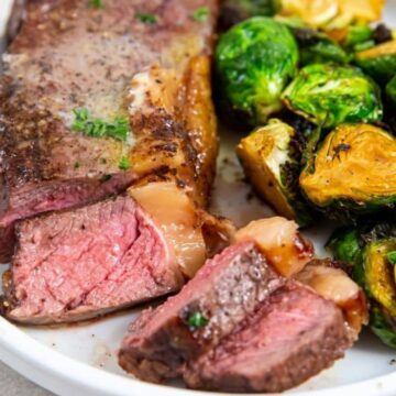 A plate with sliced medium-rare steak and a side of roasted Brussels sprouts. A fork is placed on the right side of the plate. The dish is presented on a white plate on a light-colored surface.