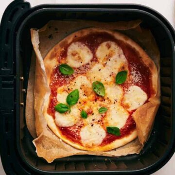 A pizza with tomato sauce, melted cheese, and fresh basil leaves is inside an air fryer on parchment paper. A jar of tomato sauce, a bowl of grated cheese, and a plate with a basil sprig are nearby on the countertop.