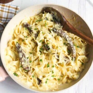 A pan filled with creamy fettuccine alfredo, a classic Italian dish, is topped with sliced mushrooms and garnished with parsley. A wooden spoon rests on the edge of the pan. A checkered cloth and parsley sprigs are visible on the white surface, creating a setting worthy of Michelin-star perfection.
