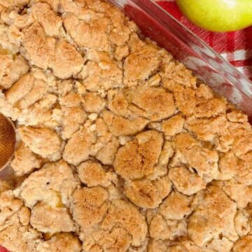 A baking dish filled with apple cobbler topped with golden-brown crumb topping. A wooden spoon rests on the left side. Two green apples are placed on a red-striped cloth next to the dish.