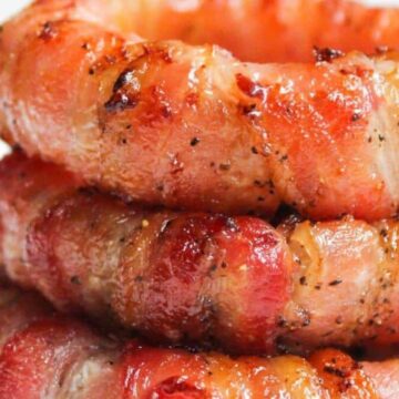 Three onion rings wrapped in crispy, grilled bacon stacked on top of each other. The bacon has a browned and peppered appearance, suggesting it is well-cooked. The background is softly blurred.