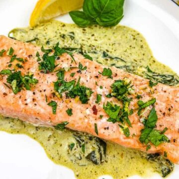 A cooked salmon fillet is topped with chopped herbs and served on a white plate. It is placed on a bed of creamy green sauce with spinach. A lemon wedge and a basil leaf are nearby, and a striped cloth is partially visible to the side.