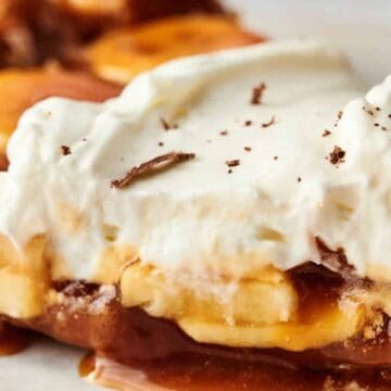 A close-up of a banoffee pie slice on a plate showcases layers of banana slices, caramel, and a crumbly crust, topped with whipped cream and chocolate shavings. Perfect for satisfying your sweet tooth, this delightful no-bake recipe is an ideal quick dessert.