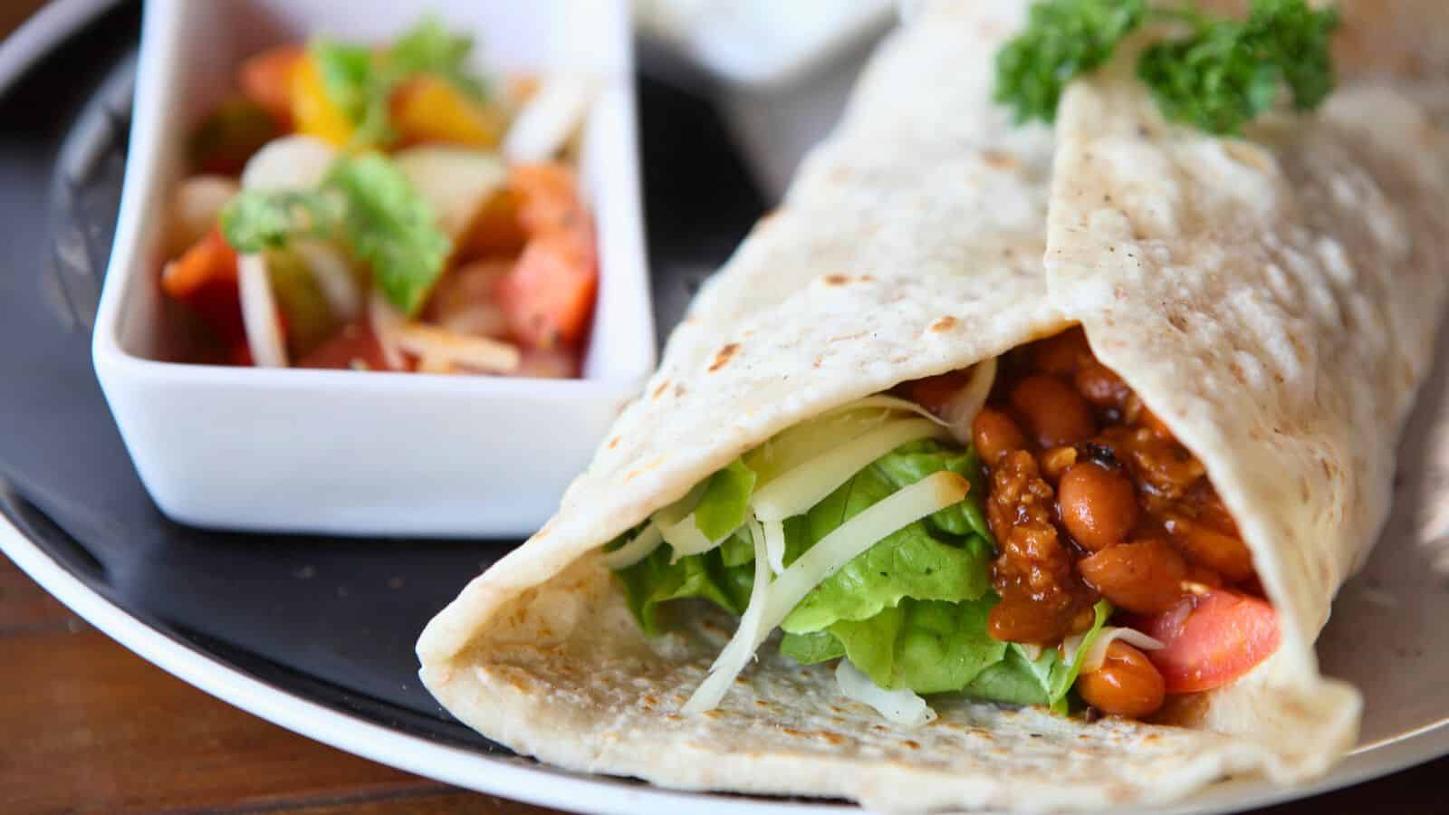 A close-up of a burrito on a plate, filled with beans, cheese, lettuce, and tomatoes. Beside it, a small rectangular dish holds a mixed salad with greens and vegetables. The plate is set on a wooden table.